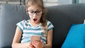 Child girl looking at mobile phone at home, with a surprise expression on her face