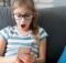 Child girl looking at mobile phone at home, with a surprise expression on her face