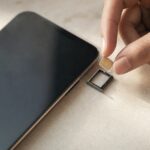 A hand holds a small SIM card beside an unbranded smartphone and its empty SIM tray.