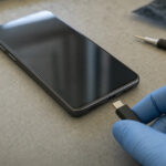 A blue-gloved hand holds a USB-C connector beside an unbranded smartphone on a device-examination mat, illustrating mobile spyware research.
